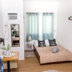 Interior view of a clean, cozy studio apartment room at MC's Place in Lapu-Lapu City. A double bed with decorative black pillows (featuring a circular G-pattern), light blue curtains, and wood-patterned flooring is visible. Potted plants on floating shelves and side tables, a wall-mounted TV, and a window AC unit complete the scene.