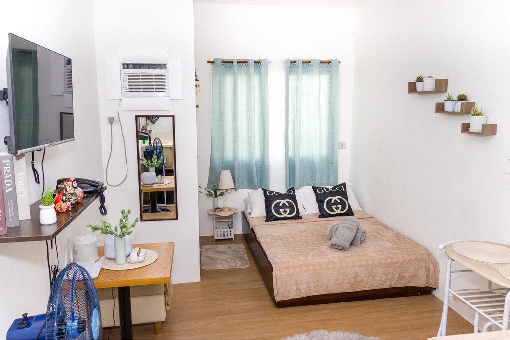 Interior view of a clean, cozy studio apartment room at MC's Place in Lapu-Lapu City. A double bed with decorative black pillows (featuring a circular G-pattern), light blue curtains, and wood-patterned flooring is visible. Potted plants on floating shelves and side tables, a wall-mounted TV, and a window AC unit complete the scene.