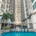 An exterior view of a clear blue outdoor swimming pool at The A. Venue Hotel in Manila, Philippines. Two large palm trees are on the left, and a multi-story building with white louvered facades frames the pool area on the right, all under a bright sky. The text "The A. Venue Hotel, Manila, Philippines" is visible on a stone-block divider element.