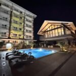 Nighttime view of an outdoor swimming pool with black lounge chairs next to a multi-story apartment building and an illuminated glass pavilion at ANIA GUEST ROOM in Cebu.