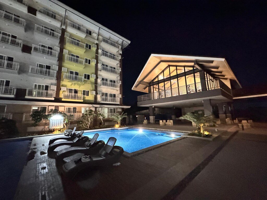 Nighttime view of an outdoor swimming pool with black lounge chairs next to a multi-story apartment building and an illuminated glass pavilion at ANIA GUEST ROOM in Cebu.