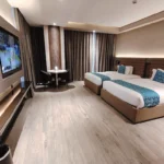Modern twin queen bedroom at Bayprime Hotel in Manila, Philippines, featuring two beds with blue accents, a wall-mounted flat-screen TV, and a small dining table.