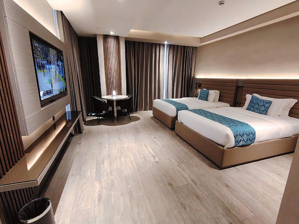 Modern twin queen bedroom at Bayprime Hotel in Manila, Philippines, featuring two beds with blue accents, a wall-mounted flat-screen TV, and a small dining table.