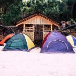 Several colorful camping tents pitched on a white sand beach, in front of traditional bamboo huts at Borawan Island Resort, Padre Burgos, Quezon, Philippines, with a lush jungle cliffside in the background.