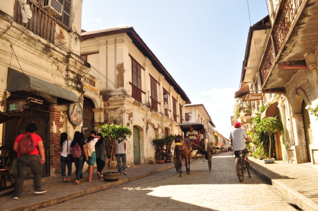 A cobblestone street lined with Spanish colonial-era houses and horse-drawn carriages under a warm twilight sky in Calle Crisologo, Vigan, Philippines.