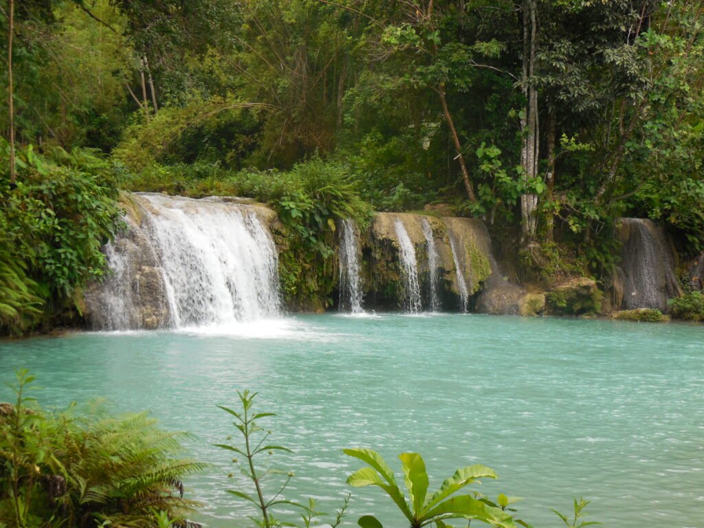 A traveler swinging from a rope vine into the tiered turquoise pools of Cambugahay Falls in Siquijor, Philippines, surrounded by lush green tropical jungle.
