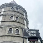 Low-angle view of Camelot Hotel in Quezon City, featuring its unique medieval castle-inspired circular stone tower facade, arched windows, and a black hotel sign under an overcast sky.