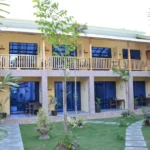 Two-story yellow building of Captain's Lodge and Bar in Bohol, featuring wooden balcony railings and a stone path winding through a green grass lawn.