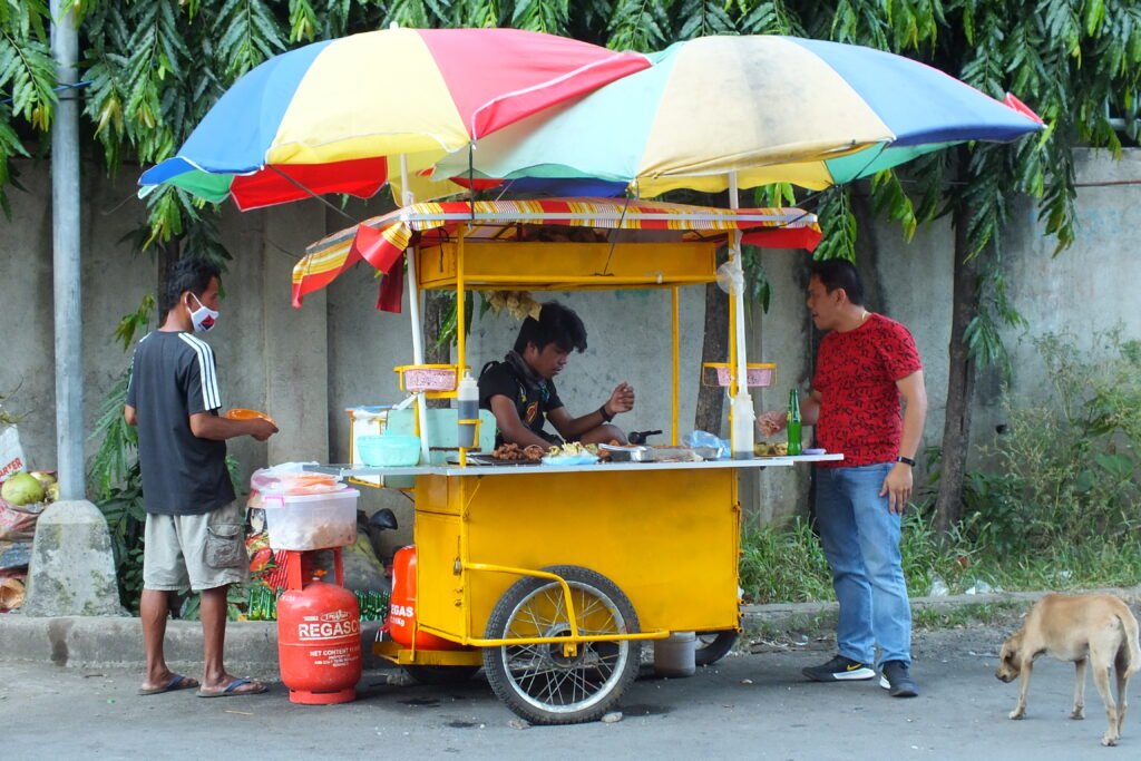 A vibrant outdoor night market stall in Cebu, Philippines, showcasing popular Cebuano street foods including sticks of grilled pork barbecue, ginabot (crispy chicharon bulaklak), and puso (hanging rice) wrapped in coconut leaves.