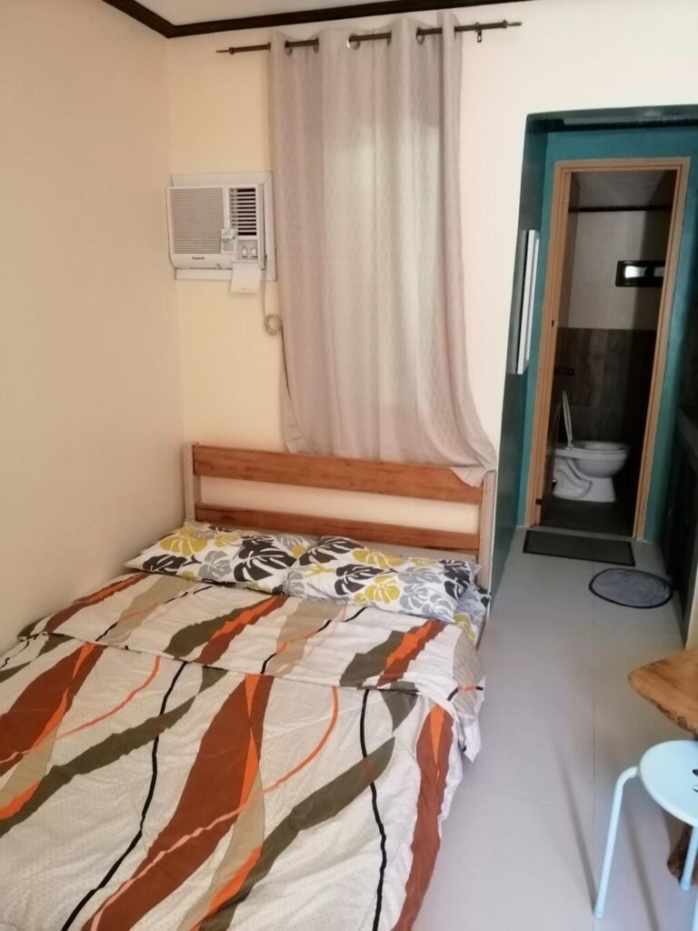 Interior view of a private guest room at CK Homestay Siargao, showing a double bed with patterned sheets, wall air conditioner, and an open bathroom door.