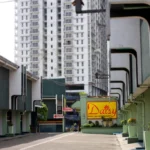 Exterior driveway view of Daisy Drive-Inn & Restaurant, showing multi-level garage units with green and white architecture, a bright yellow sign, and a tall modern apartment building in the background.