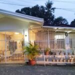 A high-definition photograph of a modern Filipino bungalow with a new, stone-coping illuminated plunge pool and a waterfall feature. The plaque on the pool reads 'PUAN MINI POOL OASIS, DAVAO CITY'. The scene is set at dusk with warm exterior and interior lights and updated solar path lights.
