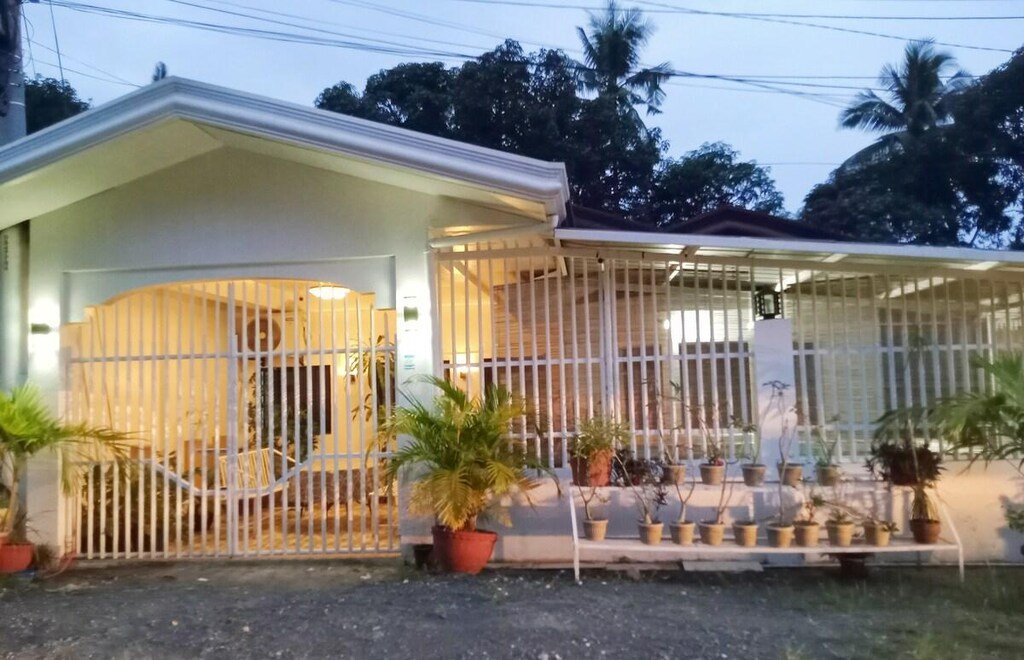 A high-definition photograph of a modern Filipino bungalow with a new, stone-coping illuminated plunge pool and a waterfall feature. The plaque on the pool reads 'PUAN MINI POOL OASIS, DAVAO CITY'. The scene is set at dusk with warm exterior and interior lights and updated solar path lights.
