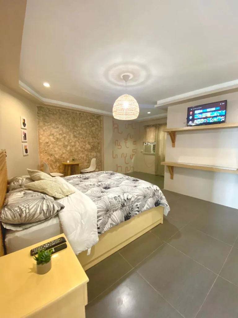 Interior view of Estancia Boho Chic Room 1 in Cebu, Philippines, featuring a bed with a black and white tropical palm leaf duvet, a woven rattan pendant light, a textured accent wall, and wall-mounted wooden shelves beneath a flat-screen TV.