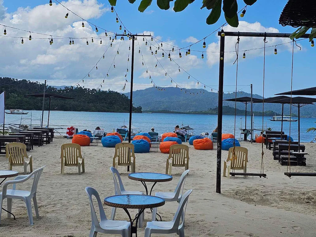 Outdoor beachfront lounge area at Ferellee Beachfront Inn in Palawan, featuring colorful bean bags, tables, and swings on the sand overlooking the ocean.