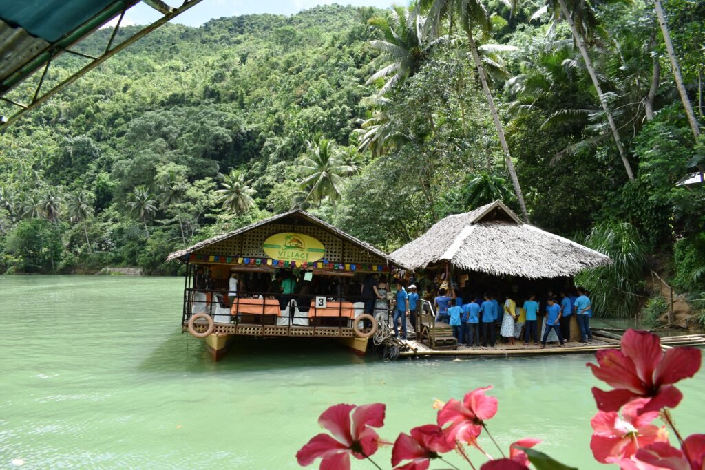 A traditional wooden floating restaurant boat cruising along the lush, emerald-green waters of the Loboc River in Bohol, Philippines, surrounded by tropical palm trees.