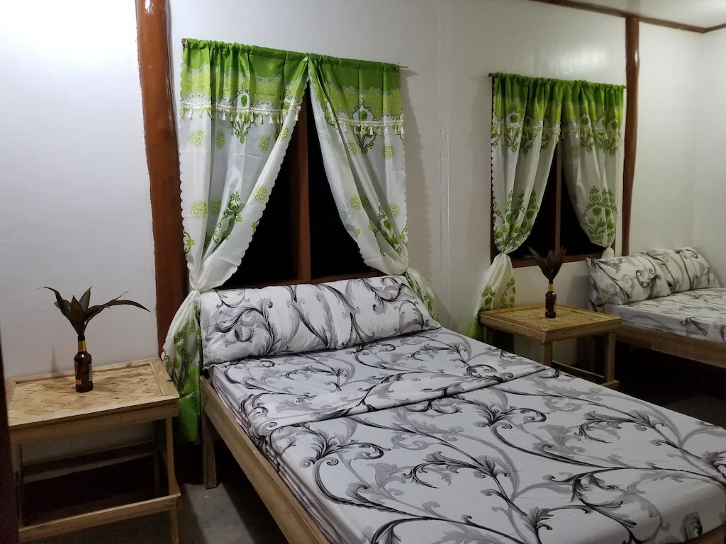 Interior view of a guest room at FN-Lar's Transient in Palawan, Philippines, featuring two beds with black-and-white patterned linens, bamboo side tables, and green-and-white decorative curtains.