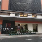 The modern multi-story exterior facade and main street-level entrance of G Square Residences in Manila, Philippines, featuring contemporary architecture, terracotta accents, horizontal louvers, a black sign with a red 'G' logo, and a row of potted palms along the sidewalk.