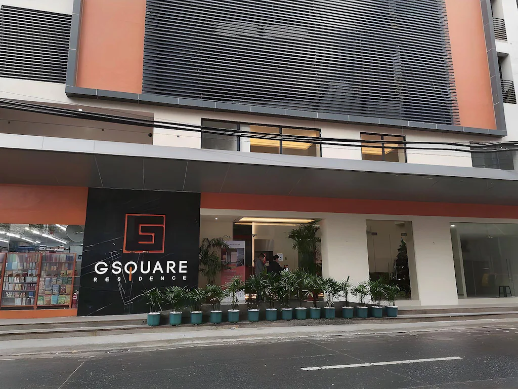 The modern multi-story exterior facade and main street-level entrance of G Square Residences in Manila, Philippines, featuring contemporary architecture, terracotta accents, horizontal louvers, a black sign with a red 'G' logo, and a row of potted palms along the sidewalk.