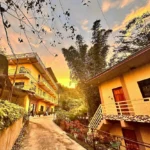 Exterior view of Garden nen Ines Guest House in La Trinidad, Benguet, Philippines, featuring multi-story yellow buildings, a paved driveway, and lush greenery under a golden sunset sky.