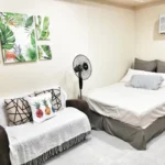 A homey studio condo unit beside SM Uptown, Cagayan De Oro, Philippines, featuring a white bed with gray pillows, a sofa with pineapple throw pillows, tropical leaf wall art, an electric fan, and a window air conditioning unit.