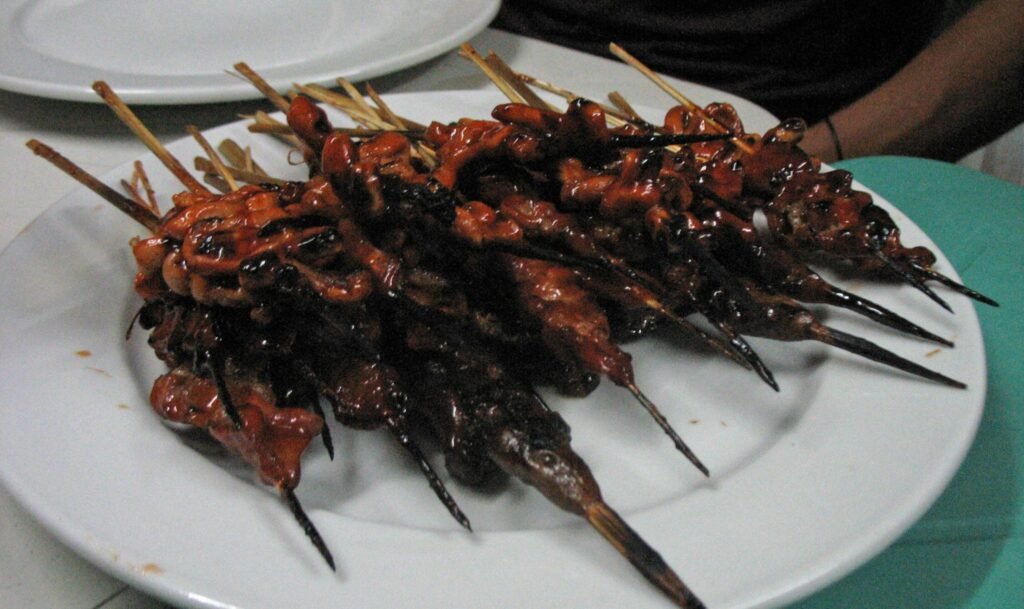 A close-up shot of freshly grilled Isaw (chicken intestines) on wooden skewers, lightly charred and glazing with sweet-spicy barbecue marinade on a smoky street food grill in Manila, Philippines.