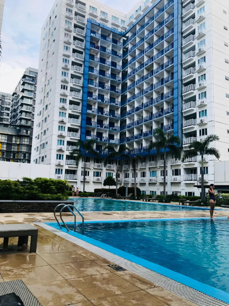Daytime view of the outdoor swimming pool area at Jamesea Residences in Manila, Philippines, featuring an expansive blue pool, poolside lounging spaces, palm trees, and a tall, modern white-and-blue residential high-rise building in the background.