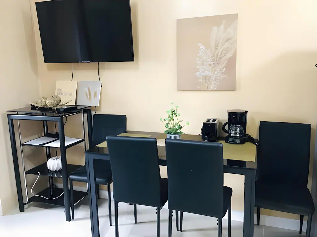 Modern compact dining area at Jazz Staycation in Manila, Philippines, featuring a black glass-top table with dark chairs, a wall-mounted TV, a toaster, and a coffee maker.