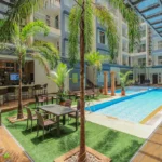 Sunlit courtyard at La Grande Residence featuring a long blue lap pool, an outdoor bar, patio dining tables, and tall palm trees under a glass awning.