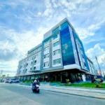 Low-angle exterior view of L'ASUNCION Hotel in Baybay City, Leyte, featuring its modern glass corner facade and a passing motorcycle under a bright, cloudy sky.