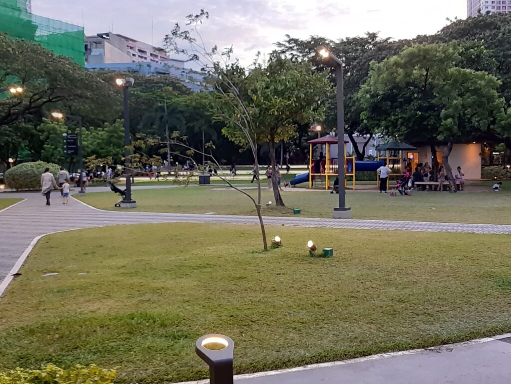 A cozy, tree-lined street in Legazpi Village, Makati, featuring trendy outdoor cafes, pedestrians walking past modern boutique buildings, and lush green canopies under bright daylight.