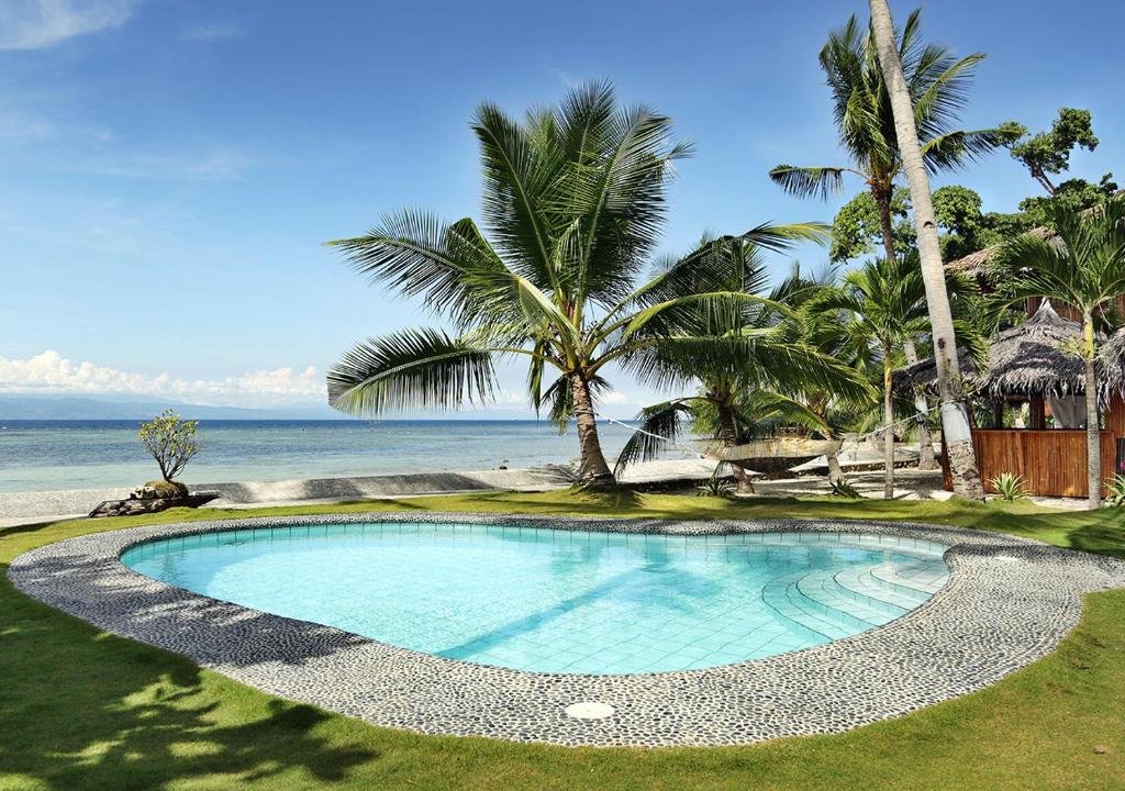 A clear, curved outdoor swimming pool lined with pebble stones on a grassy lawn at Les Maisons D' Itac resort in Cebu, overlooking a calm beach and blue ocean.