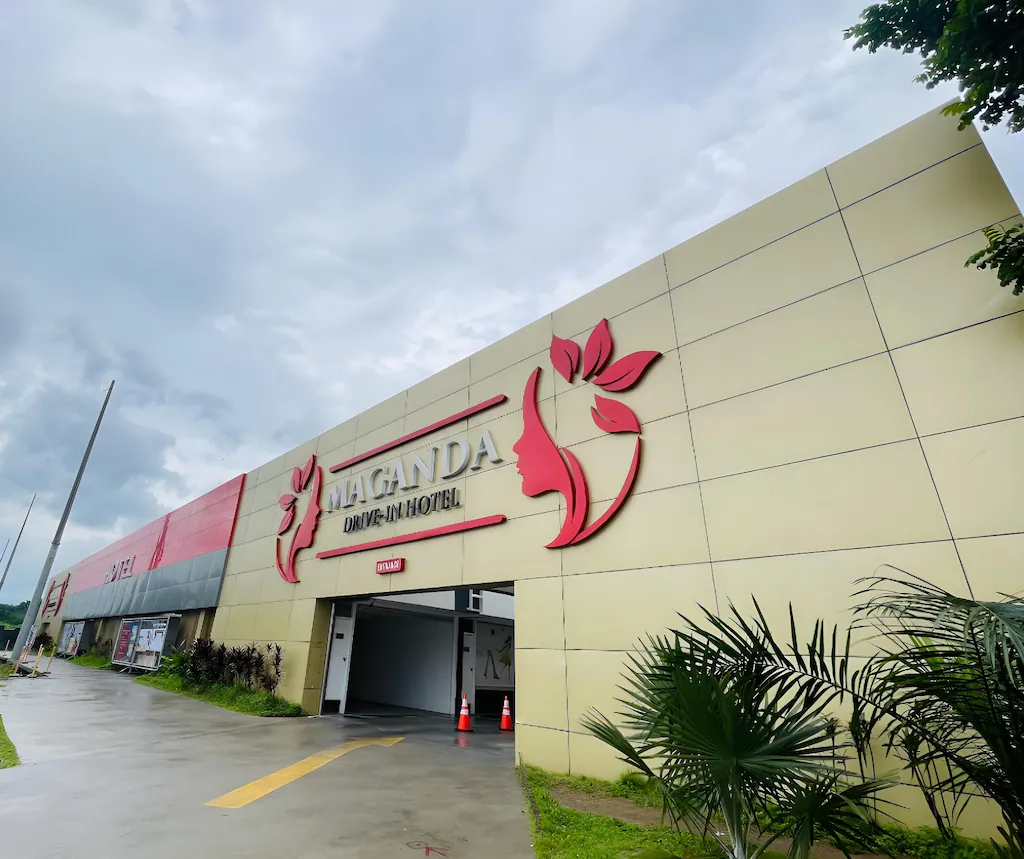 Low-angle view of the exterior entrance of Maganda Drive-In Hotel in San Fernando, Pampanga, showing a tan facade with a red logo under an overcast sky.