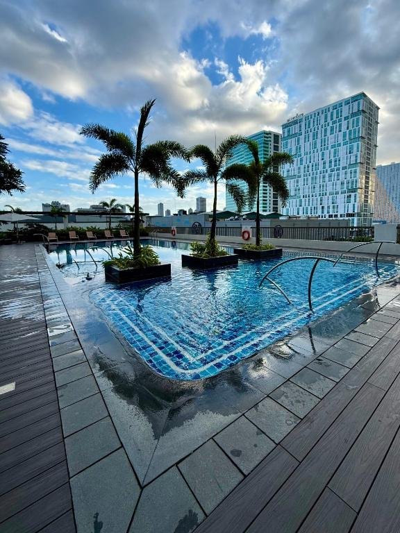 A wide-angle view of the luxurious rooftop swimming pool at Manhattan Plaza Tower 1 in Araneta City, Manila, Philippines, showing palm trees planted in the water, lounging chairs, a clear city skyline in the background, and a bright, cloudy sky, designed to look inviting for staycations.