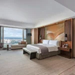Interior of a spacious luxury guest room at Marco Polo Ortigas Manila in the Philippines, featuring a king bed with an artistic gold headboard mural, warm wood paneling, a window-side chaise lounge, and floor-to-ceiling windows overlooking the city skyline.