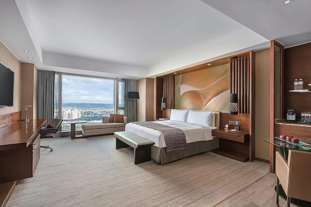 Interior of a spacious luxury guest room at Marco Polo Ortigas Manila in the Philippines, featuring a king bed with an artistic gold headboard mural, warm wood paneling, a window-side chaise lounge, and floor-to-ceiling windows overlooking the city skyline.