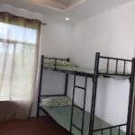 A modern bedroom inside Mia and Miguel's House in General Trias, Philippines, featuring a black metal bunk bed with green bedding, wood-look flooring, and a large curtained window.