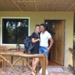 A smiling tourist couple carrying backpacks stands on the tiled veranda outside a native-style bungalow room at MIKI's Guest House on Siquijor Island.