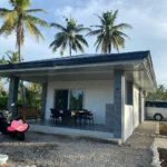 Exterior view of a modern private villa rental in Cebu, Philippines, featuring a spacious covered veranda, outdoor dining table, gravel driveway, and tall coconut palm trees under a blue sky.