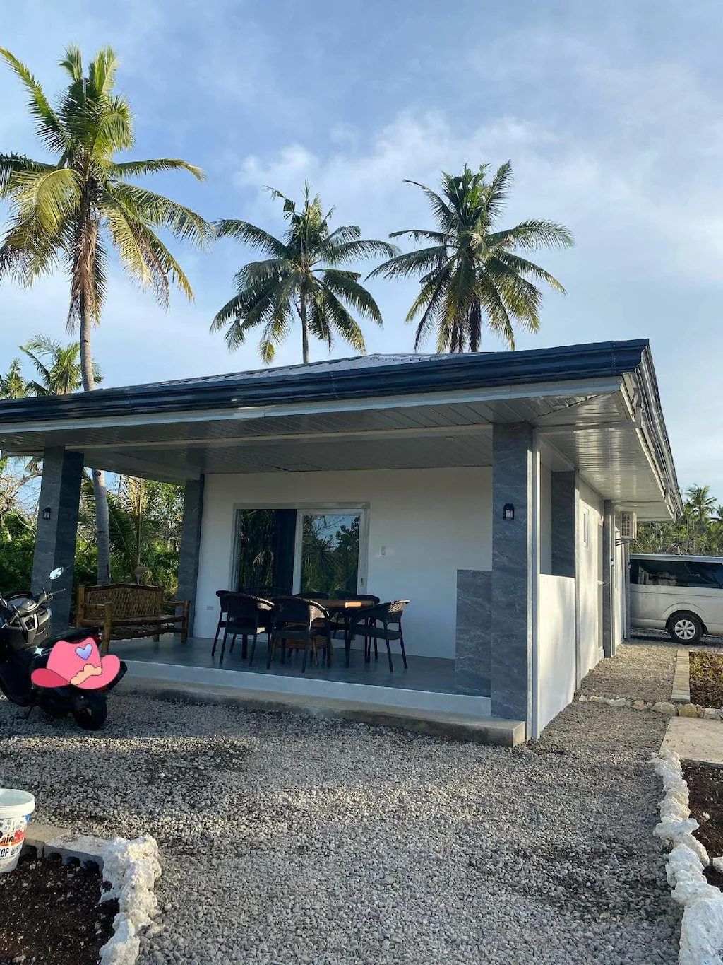 Exterior view of a modern private villa rental in Cebu, Philippines, featuring a spacious covered veranda, outdoor dining table, gravel driveway, and tall coconut palm trees under a blue sky.