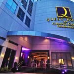 Low-angle exterior shot of the main entrance of Quest Hotel and Grand Cenia Residences in Cebu, showing illuminated gold lettering and a modern canopy facade at dusk.