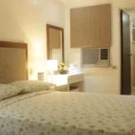 Cozy hotel room interior at Ridgecrest Gardens Hotel in Subic, featuring a bed with green palm leaf print linens and an en-suite bathroom view.