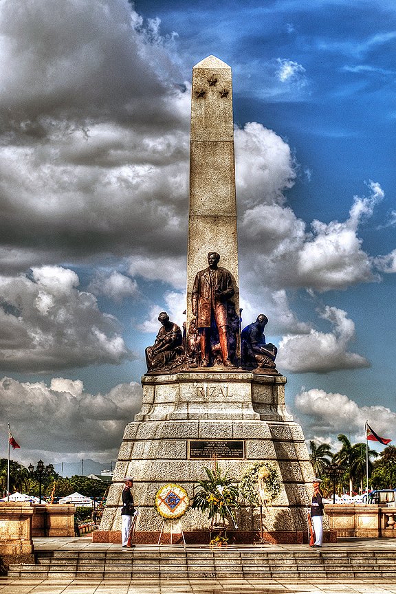 The bronze and granite monument of Dr. Jose Rizal at Rizal Park (Luneta) in Manila, Philippines, guarded by soldiers under a bright blue sky.