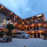 Exterior courtyard view of the three-story RSG Microhotel in General Santos City, Philippines, featuring peach-colored walls, black balcony railings, warm outdoor ceiling lights at dusk, and a silver passenger van parked on the concrete driveway.
