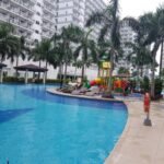 Large outdoor resort-style swimming pool surrounding a central kids' playground and lounge deck at Shell Residences in Manila, bordered by tall condo towers.