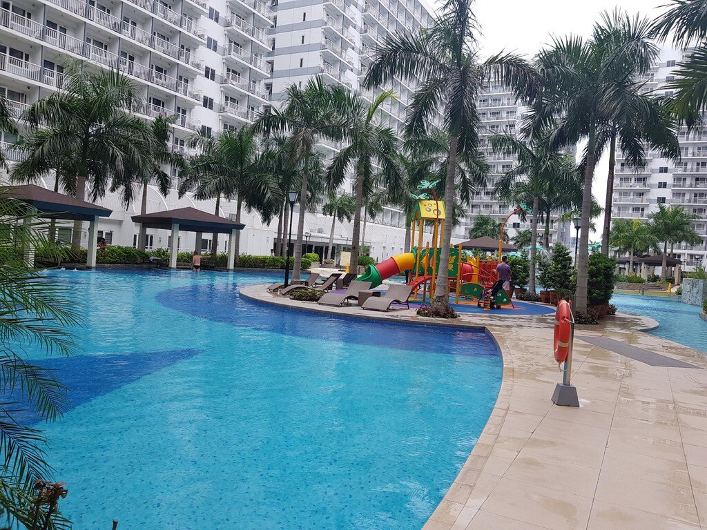 Large outdoor resort-style swimming pool surrounding a central kids' playground and lounge deck at Shell Residences in Manila, bordered by tall condo towers.