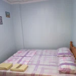 Budget-friendly guest room at Sky Beach Rooms in Cebu, Philippines, featuring a double bed with a purple and white plaid sheets set, a wooden headboard, light blue walls, and folded yellow towels.