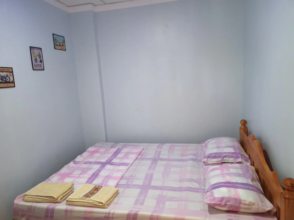 Budget-friendly guest room at Sky Beach Rooms in Cebu, Philippines, featuring a double bed with a purple and white plaid sheets set, a wooden headboard, light blue walls, and folded yellow towels.