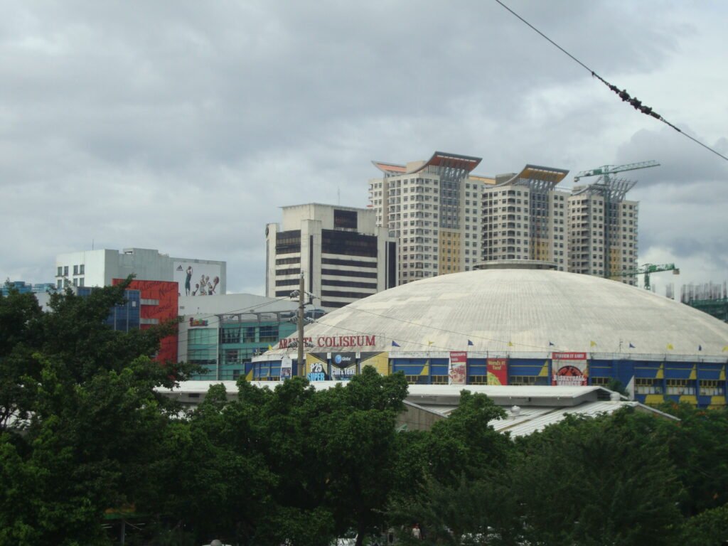 The iconic white dome of the Smart Araneta Coliseum in Quezon City, Manila, Philippines, framed by lush green trees and modern high-rise buildings under a cloudy sky.