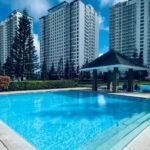 Outdoor swimming pool at Tagaytay Staycation by SMDC Wind Residences Lakeview in the Philippines, featuring a covered poolside gazebo, pine trees, and modern high-rise condo towers under a cloudy blue sky.